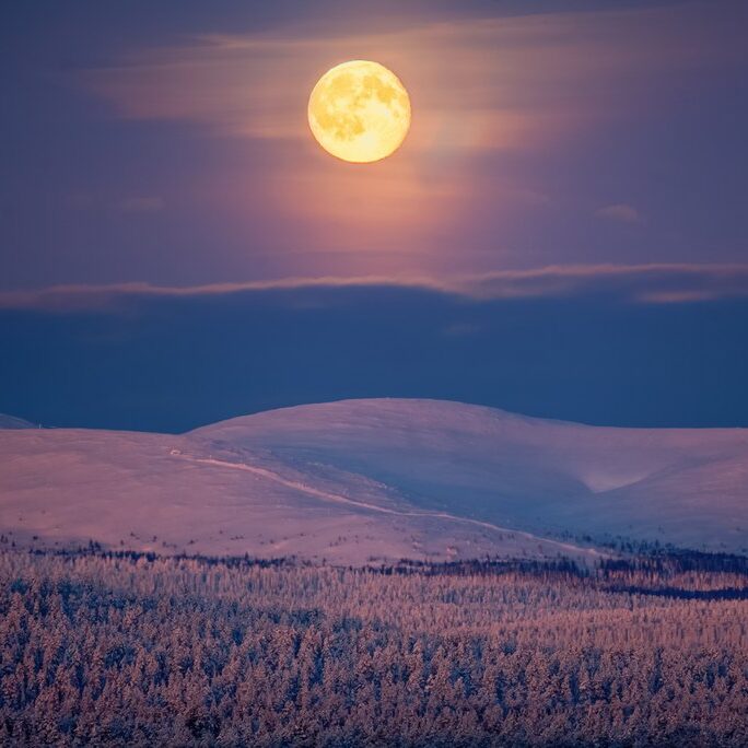 A bright full moon illuminates a snowy mountain range during the serene polar night in Muonio, Finnish Lapland