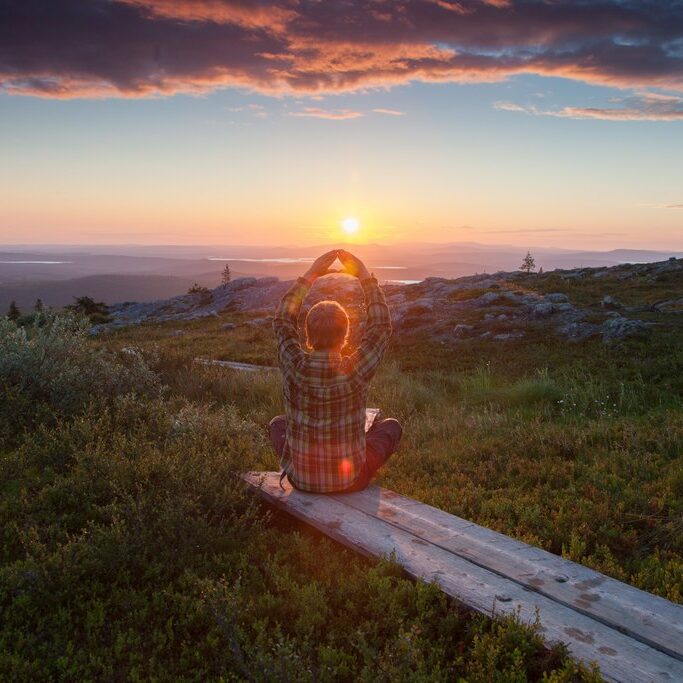 A person relaxes on a wooden path on top of a fell enjoying the warm glow of a midnight sun in the background in Muonio, Finnish Lapland
