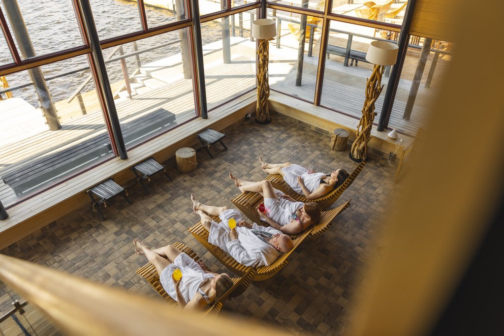 Four people relax in the sauna lounge at Arctic Sauna World – one reclines on a wooden chair wrapped in a towel, sipping water, while large windows reveal a scenic view of the lake and forest outside in Lapland, Finland
