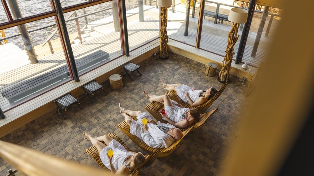 Four people relax in the sauna lounge at Arctic Sauna World – one reclines on a wooden chair wrapped in a towel, sipping water, while large windows reveal a scenic view of the lake and forest outside in Lapland, Finland