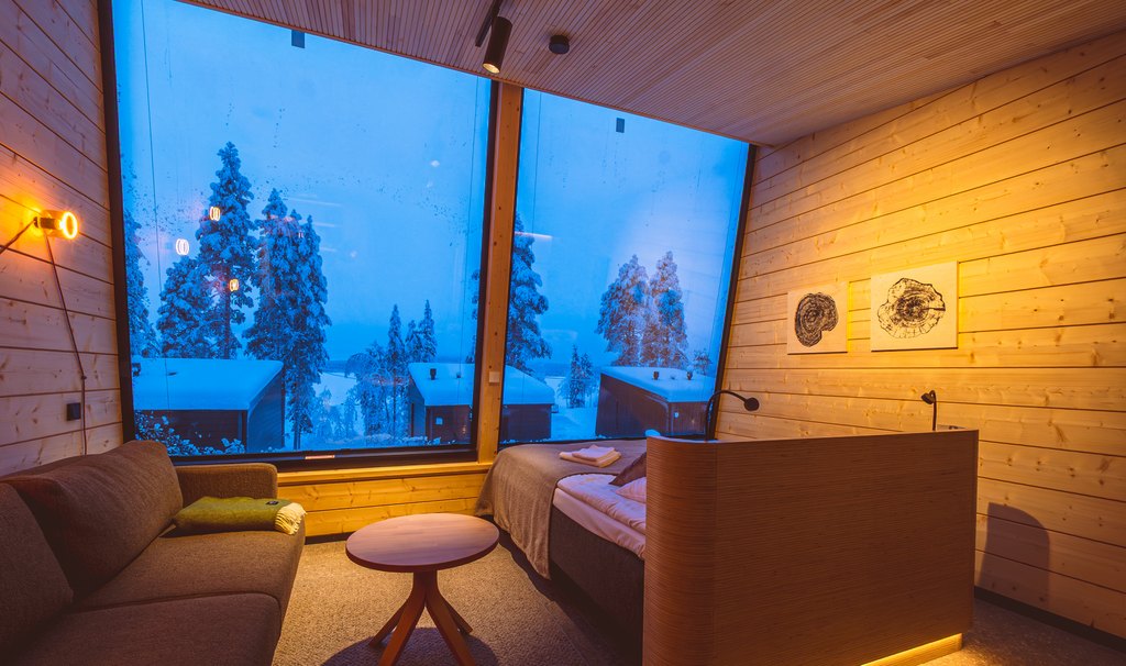 FI: Jeris Lakeside Resortin Arctic Sky Nest -huone sinisen hetken Lapissa: suuri panoraamaikkuna avautuu lumiseen metsään ja mökkimaisemaan, sisällä lämmin puusisustus, sohva ja parivuode tunnelmavalaistuksessa.
EN: Jeris Lakeside Resort Arctic Sky Nest room at blue hour in Finnish Lapland: a large panoramic window looks out to a snowy forest and cabins, with warm wood interiors, a sofa, and a double bed in cosy ambient lighting.