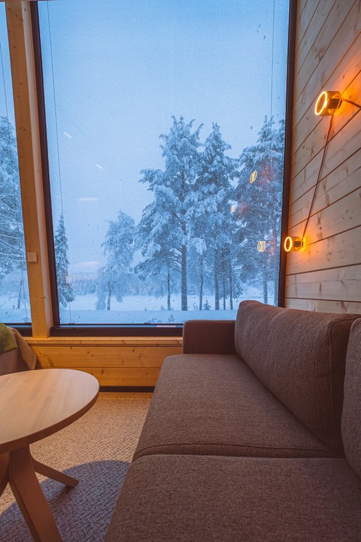 FI: Jeris Lakeside Resortin Arctic Sky Nest -oleskelutila: iso panoraamaikkuna avautuu lumiseen metsään ja järvimaisemaan, sisällä puusisustus, harmaa sohva ja pyöreä pöytä, seinällä lämmin tunnelmavalaistus.
EN: Jeris Lakeside Resort Arctic Sky Nest living area: a large panoramic window opens to snowy forest and lakeside scenery, with warm wooden interiors, a grey sofa, a round table, and cosy wall lights.