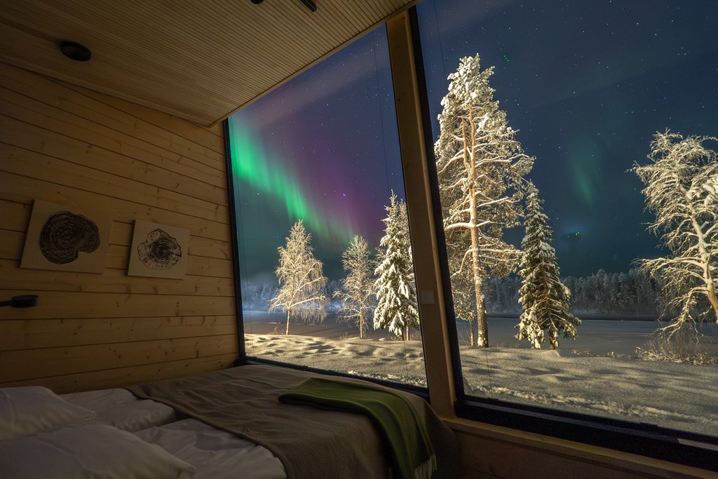 FI: Harriniva Hotels & Safarisin Harriniva Adventure Resortin Arctic Glass Suite -majoitus Muoniossa, Lapissa: pedatulta sängyltä avautuu suuri panoraamaikkuna jäätyneelle, lumen peittämälle joelle, jonka rannalla seisoo lumisia puita; tähtitaivaalla vihreät revontulet loimuavat ja lämmin puusisustus viimeistelee tunnelman. EN: Harriniva Hotels & Safaris Harriniva Adventure Resort Arctic Glass Suite in Muonio, Lapland: from the made bed, a large panoramic window looks out over a frozen, snow-covered river with snowy trees along the riverbank; green northern lights glow in the starry sky, and warm wooden interiors complete the cosy atmosphere.