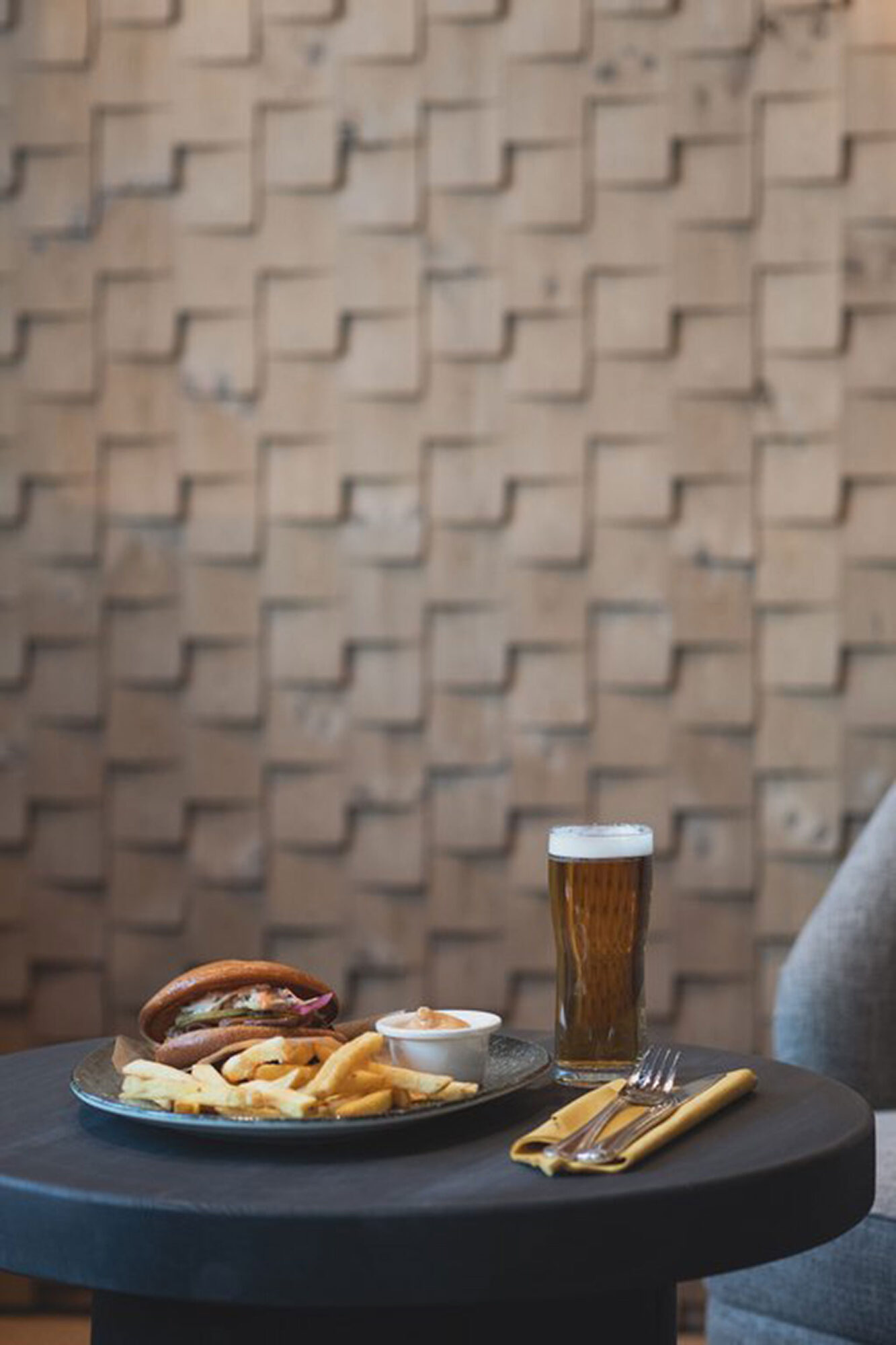 FI: Jeris Lakeside Resortin Saiho Bar & Lounge -menun annos: burgeri ja ranskalaiset dippikastikkeella pienellä pyöreällä pöydällä, vieressä lasi olutta sekä keltainen servetti ja aterimet, taustalla lämminsävyinen tiiliseinä.
EN: Dish from Jeris Lakeside Resort Saiho Bar & Lounge menu: a burger and fries with dipping sauce on a small round table, alongside a glass of beer and cutlery on a yellow napkin, with a warm-toned brick wall in the background.