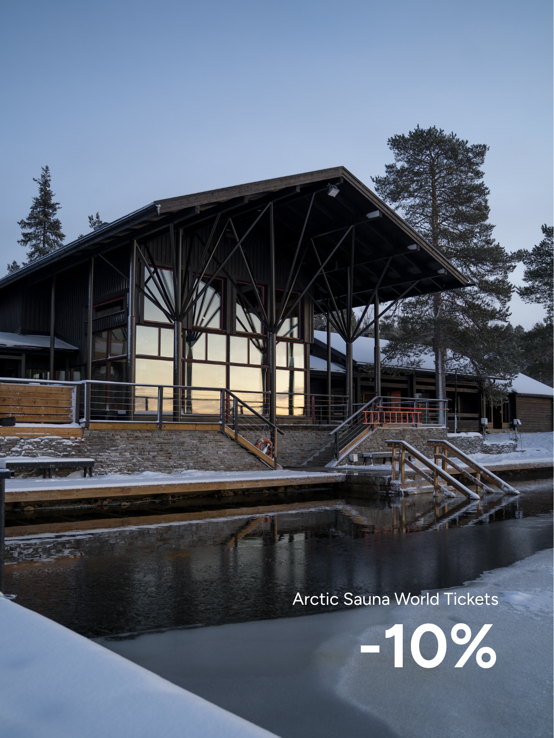 FI: Talvinen järvenrantanäkymä Jeris Lakeside Resortissa: Arctic Sauna World -saunarakennus suurine ikkunoineen heijastuu tummaan veteen, edessä avanto ja portaat, ympärillä luminen ranta ja männyt. Kuvassa teksti “Arctic Sauna World Tickets -10%” (Harriniva Hotels & Safaris).
EN: Winter lakeside view at Jeris Lakeside Resort: the Arctic Sauna World sauna building with large windows reflects on the dark water, with an ice hole and steps in the foreground, snow-covered shore and pines around. Overlay text reads “Arctic Sauna World Tickets -10%” (Harriniva Hotels & Safaris).