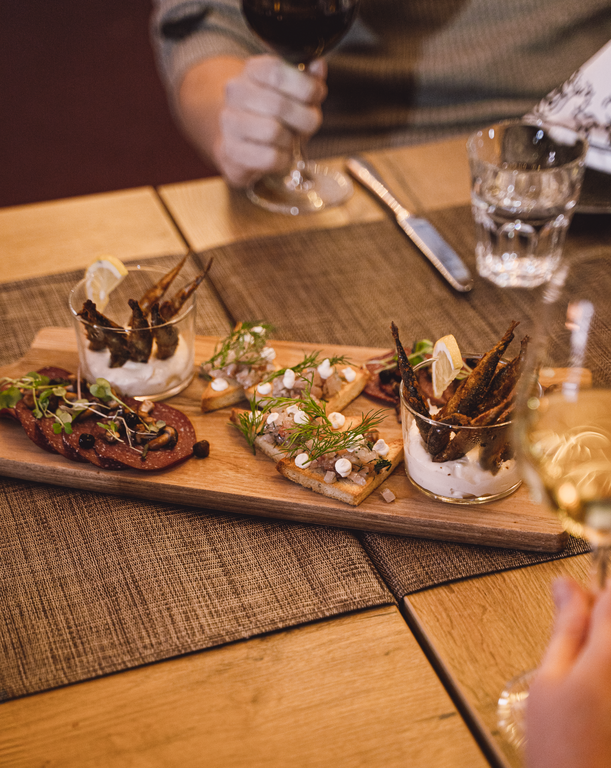 FI: Harriniva Hotels & Safarisin ja Jeris Lakeside Resortin Jierra-ravintolan Harriniva Signature Dinner Experience: jaettava alkupalalauta kynttilänvalossa—siian/lohen suupaloja, ruisleipäpaloja täytteillä, yrttejä ja rapeita pikkukaloja dipissä; ympärillä viini- ja vesilasit sekä lämmin ravintolatunnelma. EN: Harriniva Hotels & Safaris and Jeris Lakeside Resort Jierra restaurant Harriniva Signature Dinner Experience: a share-style starter board in candlelight—bite-sized fish, rye crisps with toppings, fresh herbs, and crispy small fish in dip; wine and water glasses on the table in a warm, cosy restaurant setting.EN: Share-style starter board in candlelight: bite-sized fish, rye crisps with toppings, fresh herbs, and crispy small fish in dip; wine and water glasses on the table in a warm, cosy restaurant setting.