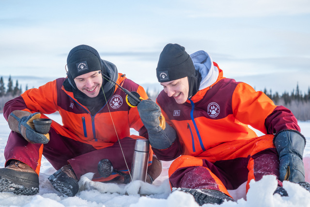 FI: Kaksi talvivaatteisiin pukeutunutta vierasta istuu lumella ja nauraa pilkkiessä jäällä; avannossa näkyy siima ja termos kuumaa juomaa – Ice fishing experience osana All-Inclusive Authentic Lapland Adventure -lomapakettia Harriniva Hotels & Safarisissa, Muoniossa. EN: Two guests in warm winter gear sit on the snow laughing while ice fishing; a fishing line drops into the ice hole beside a thermos of hot drink—an Ice fishing experience as part of the All-Inclusive Authentic Lapland Adventure holiday package with Harriniva Hotels & Safaris in Muonio, Finnish Lapland.