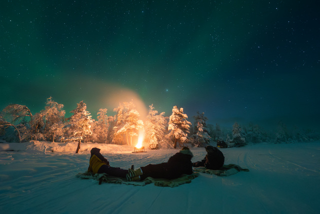 FI: Revontulet hehkuvat vihreinä tähtitaivaan alla, kun ihmiset rentoutuvat talvisella nuotiopaikalla lumisten puiden keskellä Muoniossa – All-Inclusive Authentic Lapland Adventure -lomapaketti Harriniva Hotels & Safarisissa, aitoa Lapin iltaelämystä.
EN: Northern lights glow green beneath a starry sky as guests relax by a campfire in the snowy forest in Muonio—an All-Inclusive Authentic Lapland Adventure holiday package with Harriniva Hotels & Safaris, capturing an authentic Finnish Lapland evening experience.
