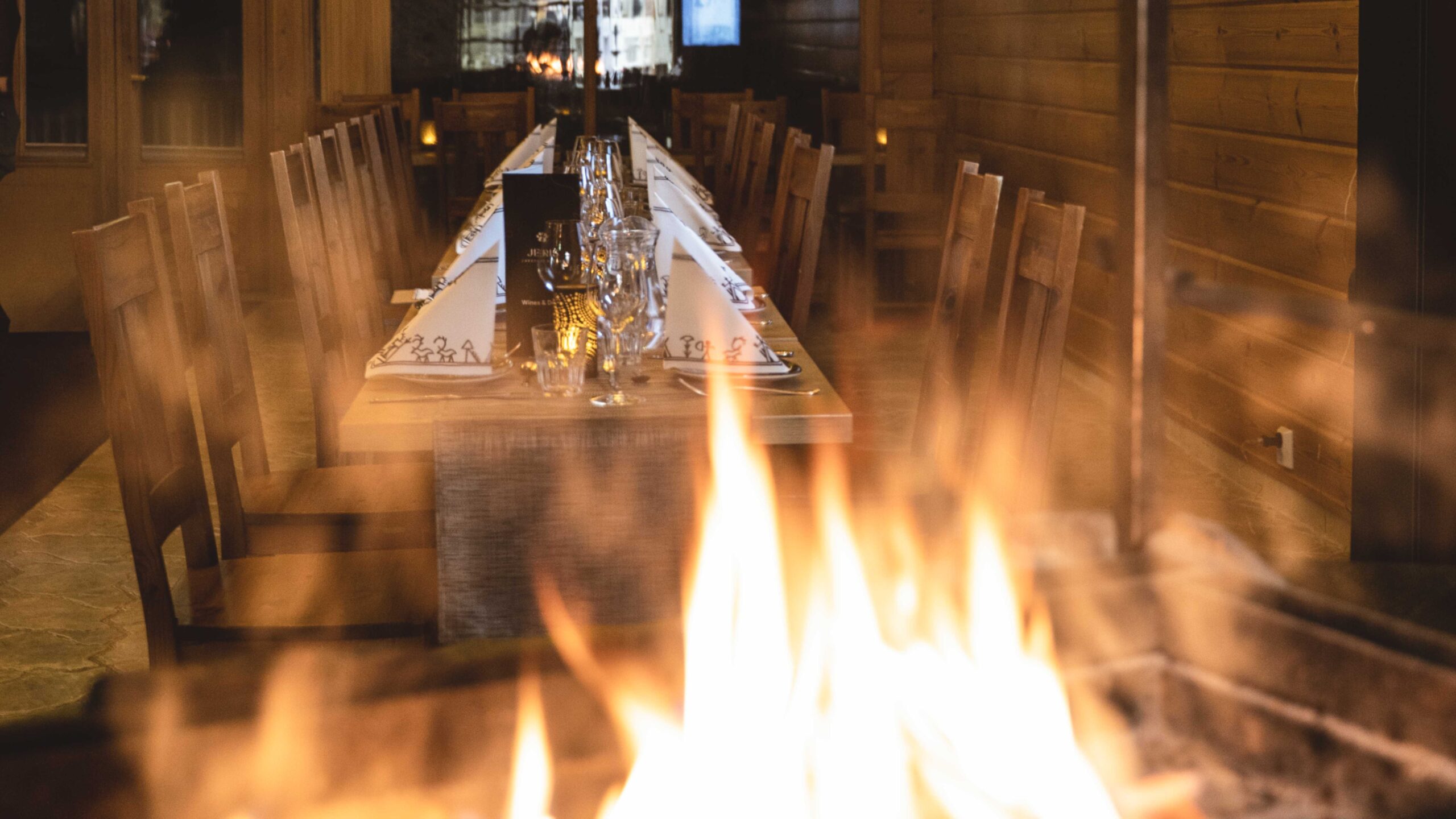 FI: Lämminhenkinen hirsiravintola ja pitkään pöytään katettu illallinen; takkatulen liekit hehkuvat etualalla ja kynttilävalo luo tunnelmaa – Harriniva Signature Dinner Experience osana Harriniva Signatures Arctic All-Inclusive -lomapakettia (Harriniva Arctic Deluxe) Muoniossa, Lapissa, Harriniva Hotels & Safaris.
EN: A cozy log-restaurant dining room with a long table set for dinner; fireplace flames glow in the foreground and candlelight creates a warm atmosphere—Harriniva Signature Dinner Experience as part of the Harriniva Signatures Arctic All-Inclusive holiday package (Harriniva Arctic Deluxe) in Muonio, Lapland, with Harriniva Hotels & Safaris.