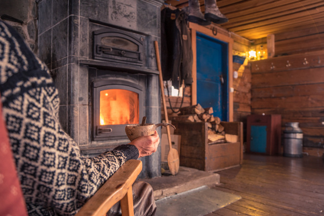 Henkilö rentoutuu takkatulen ääressä perinteisessä mökissä yöpymiselämyksen aikana Muoniossa osana Harriniva Hotels & Safarisin kesäaktiviteetteja Lapissa. Kuvassa näkyy lämmin ja kodikas mökin sisustus, puulattia, halkoja ja tunnelmallinen tulisija. A guest relaxes by the fireplace in a traditional cottage during the Overnight Cottage Experience in Muonio, part of Harriniva Hotels & Safaris summer activities in Lapland. The image shows a warm and cozy interior with wooden floors, firewood, and an atmospheric wood-burning stove.