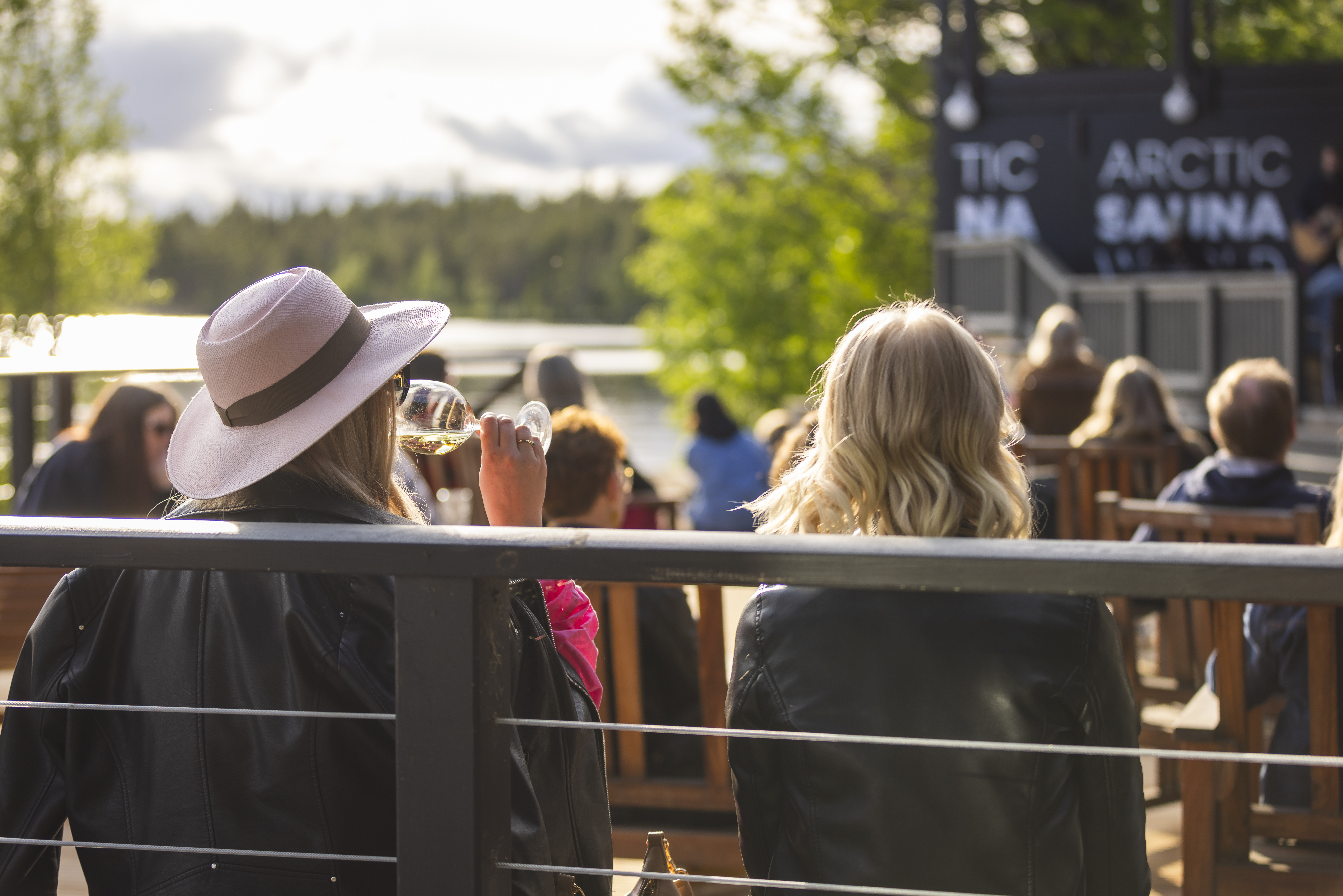Woman enjoying a glass of wine on an outdoor terrace at Arctic Sauna World in Lapland during golden hour, overlooking a lakeside setting in summer.